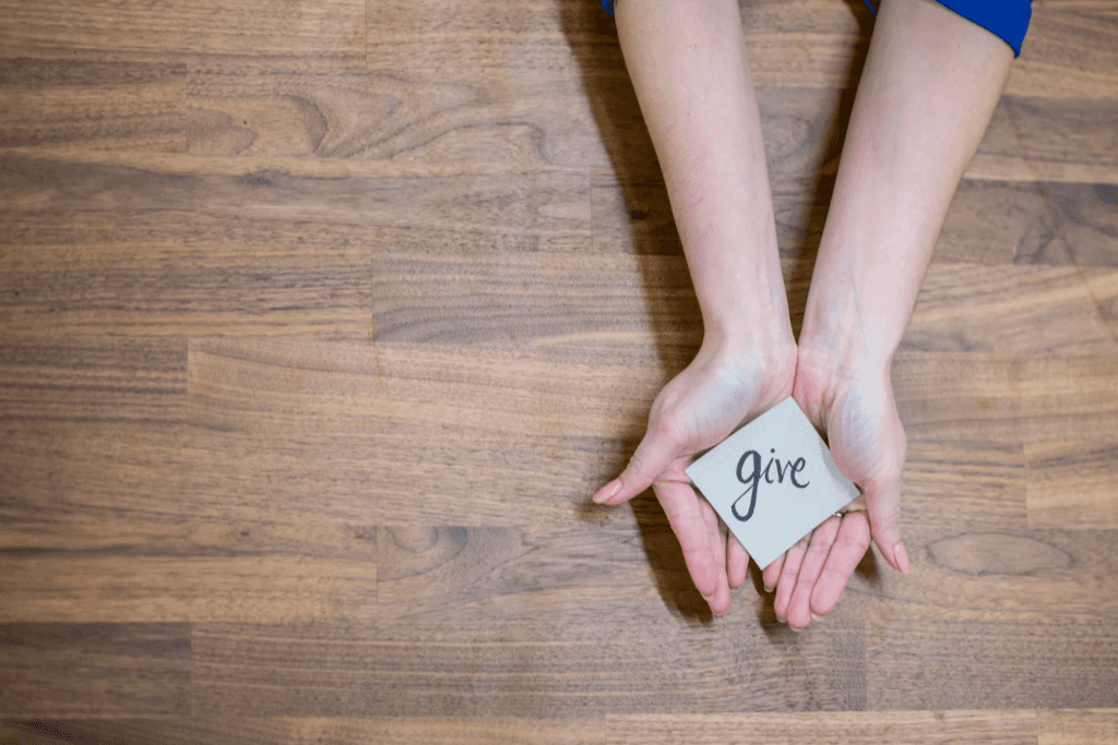 Đôi bàn tay mở ra trên nền gỗ, nâng niu một viên đá có khắc chữ "give", thể hiện sức mạnh của lòng nhân ái và tinh thần cho đi.